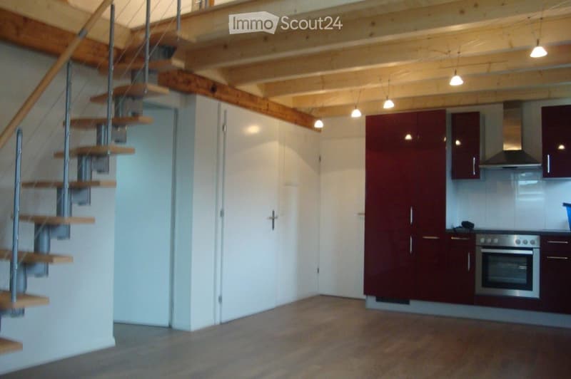 Open-plan area featuring a wooden staircase leading upwards, a modern kitchen with dark red cabinets, stainless steel oven, and range hood, and exposed wooden beams on the ceiling.