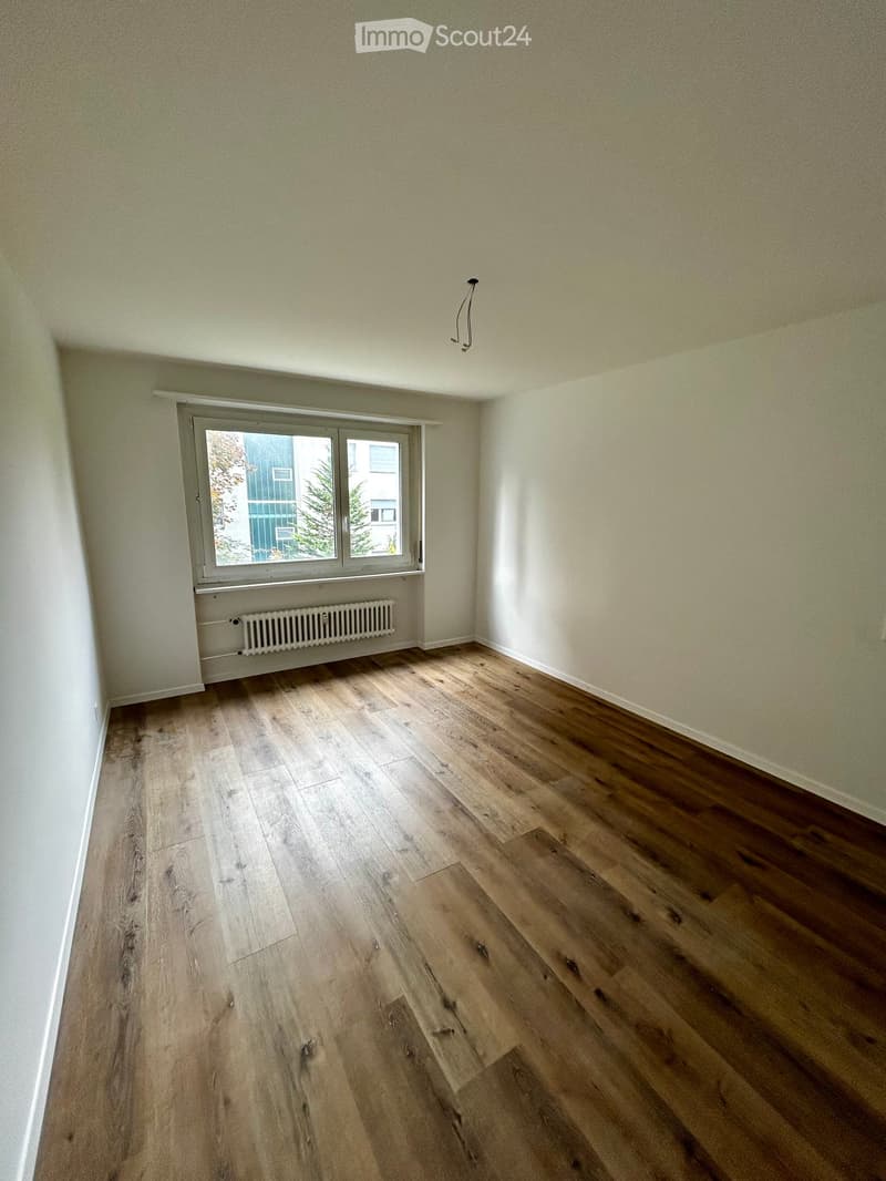 The image shows an empty room with a wooden floor, white walls, a large window, and a white heater.