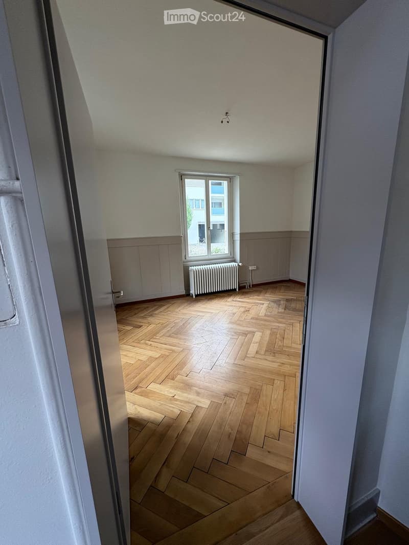 Empty room with wooden herringbone flooring, white walls, a window with a radiator below, and a visible door frame.