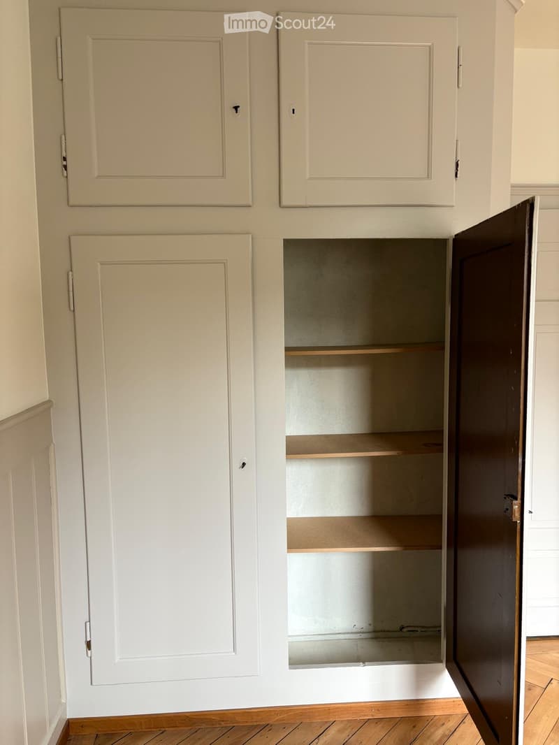 Empty kitchen cabinet with wooden shelves, white walls, and wooden flooring.