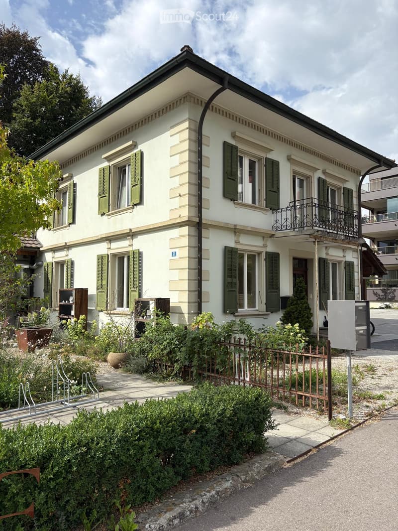 Two-story house with white facade, green shutters, and a balcony on the upper floor. The house features a well-maintained garden with shrubs and a metal fence.