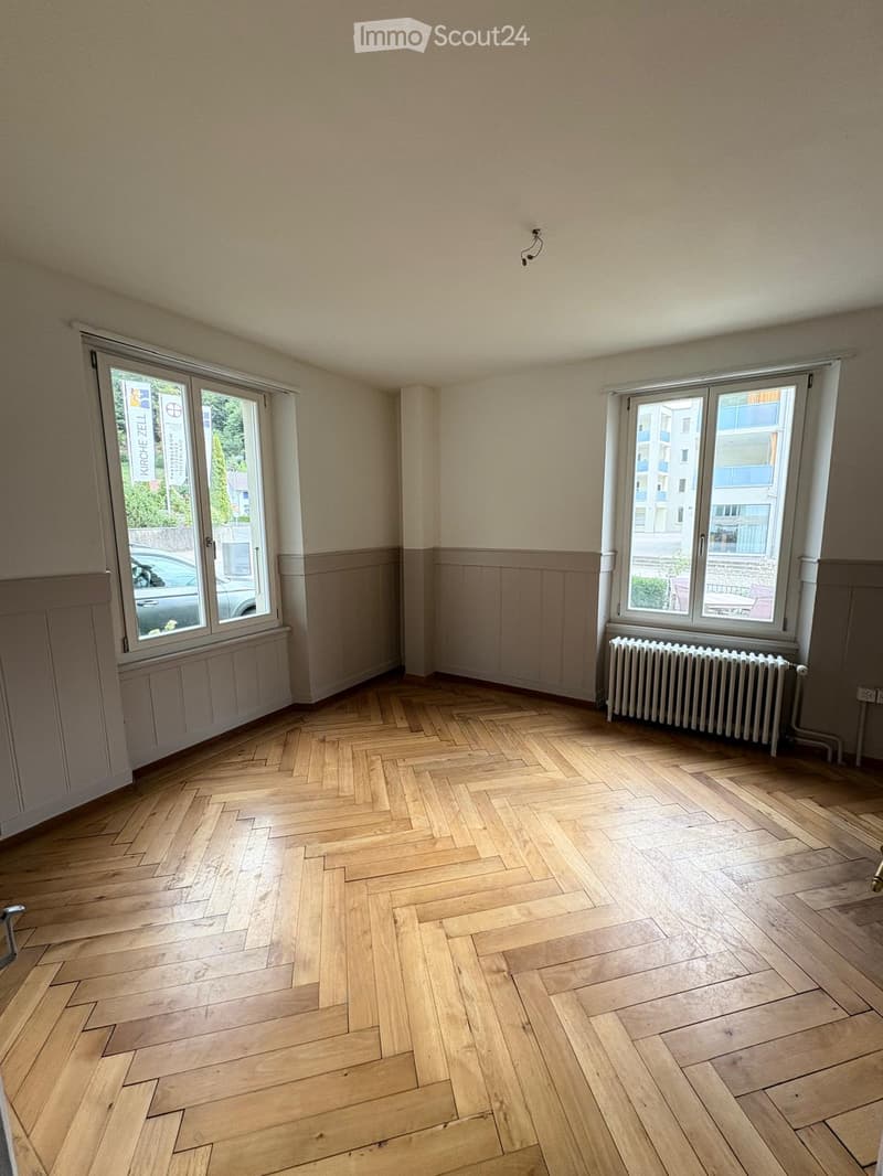 Empty room with wooden herringbone flooring, light-colored walls, and large windows with radiators beneath them.