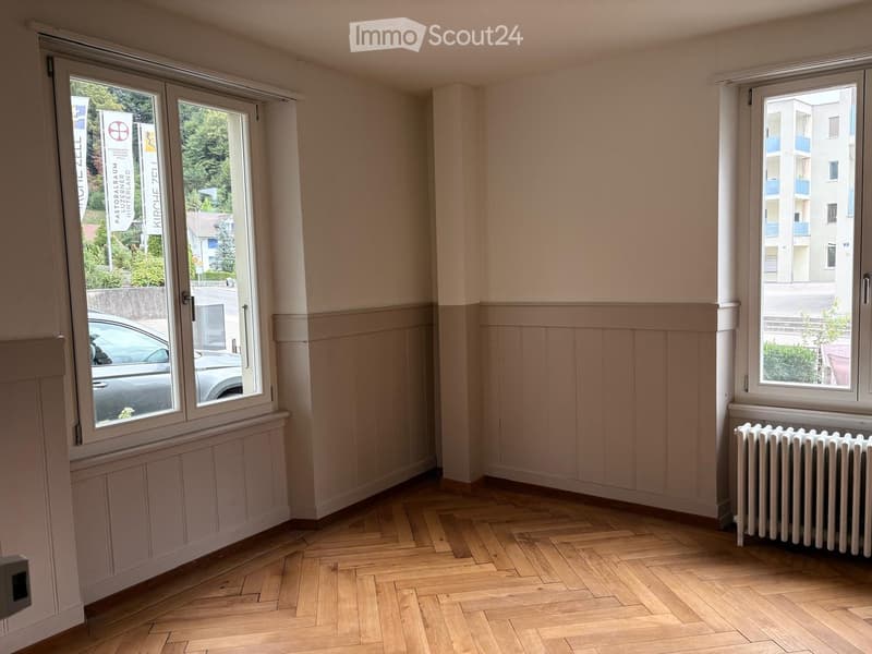 Empty room with wooden flooring, light-colored walls, and large windows offering views of the street and neighboring buildings.