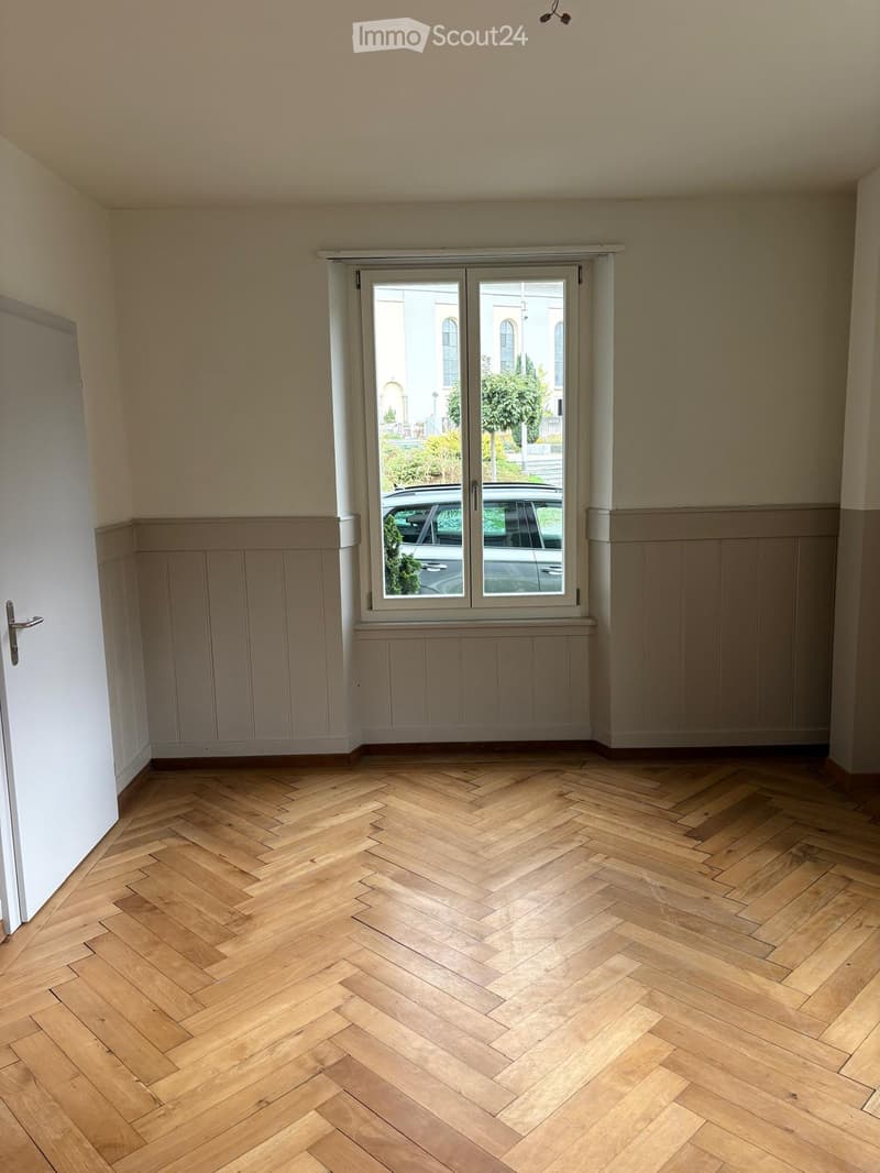 Empty room with herringbone wooden flooring, light-colored walls, and a large window with a view of a parked car and greenery outside.
