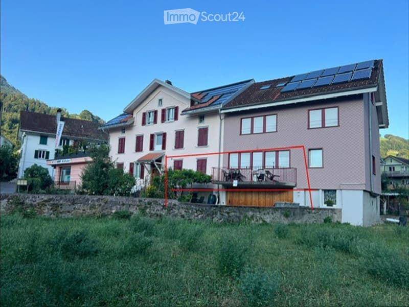 Two-story residential house with solar panels on the roof, a balcony with outdoor furniture, and a stone retaining wall in front.