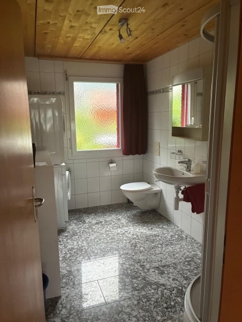 Bathroom with white tiled walls, granite floor, toilet, sink, mirror, and shower area.