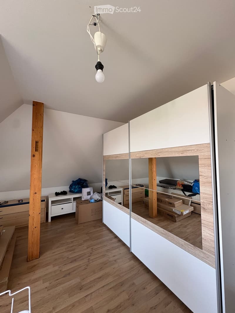A small, modern room with wooden flooring, a built-in loft bed, a desk, and storage units. The room features a hanging light fixture and a mirror on the wall.