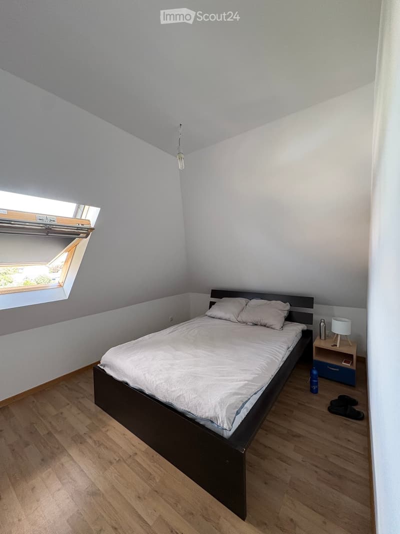 A small bedroom with a double bed, nightstand, lamp, and a window with blinds. The room features wooden flooring and white walls.