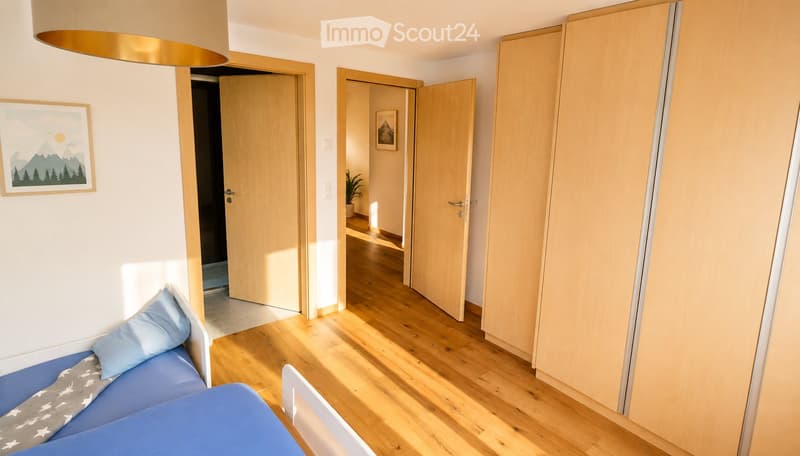 Bright bedroom with wooden flooring, a blue bed with pillows and a blanket, a wardrobe, and a picture frame on the wall. Open doors lead to other rooms.