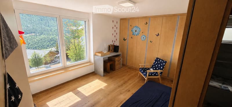 Cozy, well-lit room with wooden flooring, large windows offering a scenic mountain view, a bed, a chair, a desk, and a storage unit.