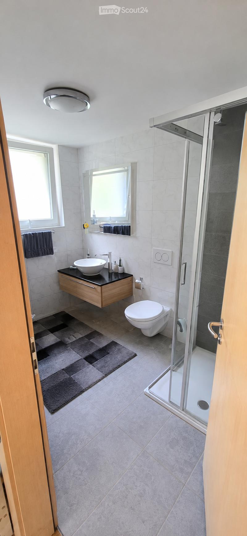 Modern bathroom with a white sink, toilet, shower, and mirror. Features a wooden vanity, dark countertop, and a grey bath mat.