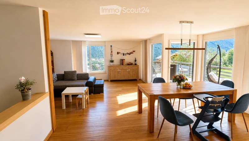 Bright living and dining area with wooden flooring, large windows offering a scenic view, a grey sofa, a coffee table, a dining table with chairs, a cabinet, and decorative plants.