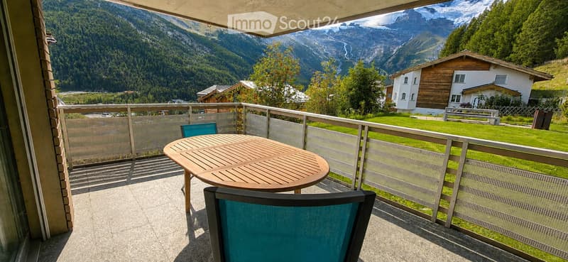 Balcony with a wooden table and two chairs, overlooking a scenic mountain landscape with houses and greenery.