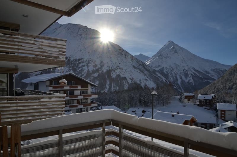 Balcony with snow-covered railing, overlooking a snow-covered mountain range and village houses under a clear blue sky with the sun shining brightly.