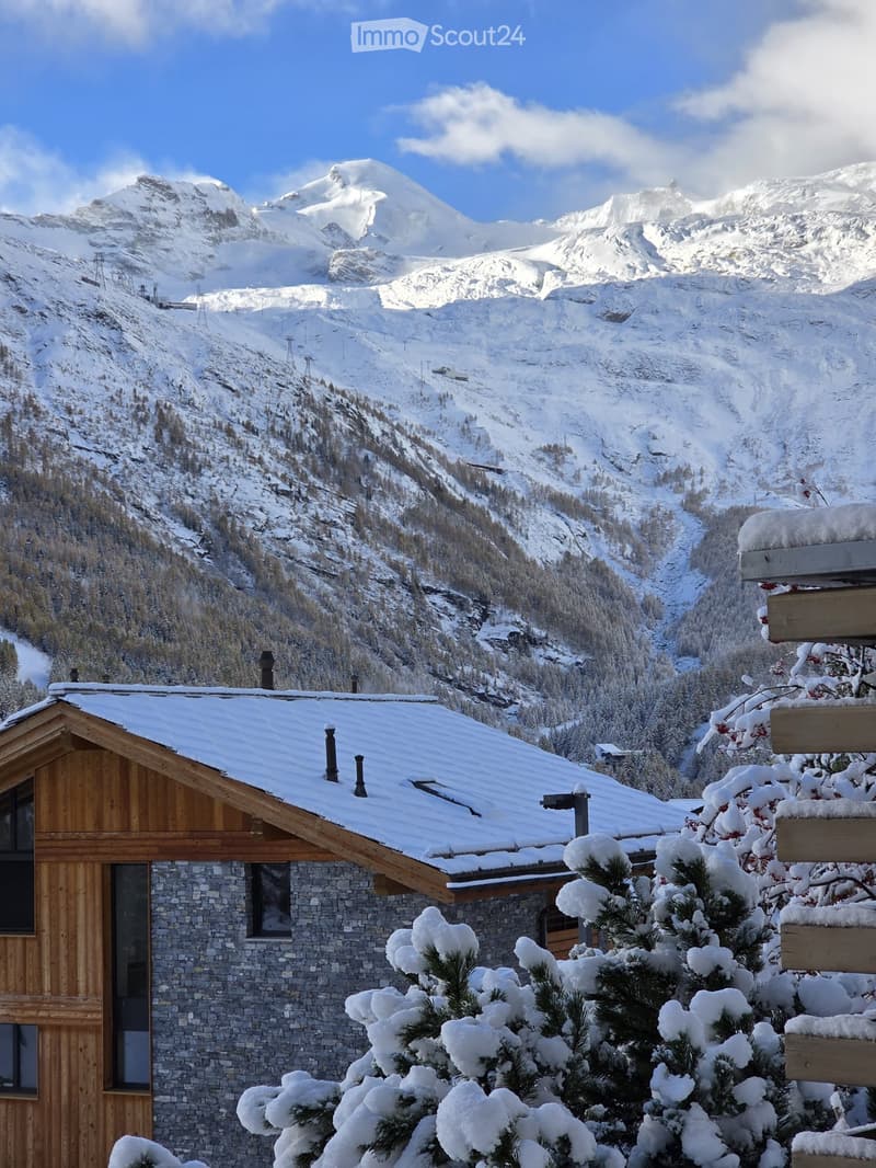 A stone and wooden house with snow-covered roof and surrounding trees, set against a backdrop of snow-capped mountains.