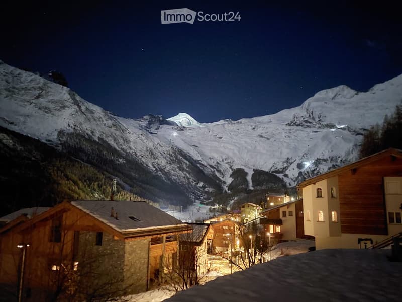 Nighttime view of a snow-covered mountain range with illuminated wooden houses and a clear starry sky.