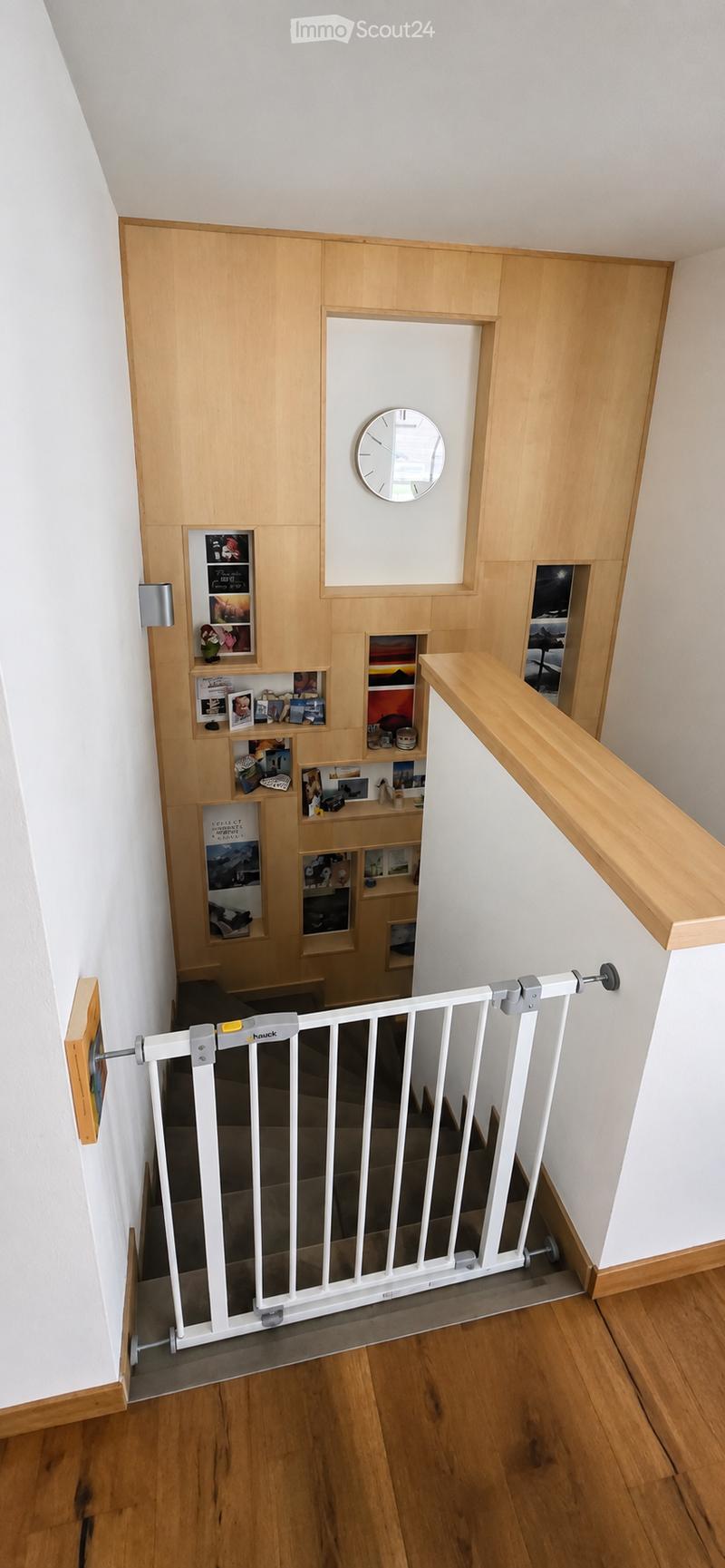 Staircase with wooden railing, white baby gate, built-in shelving unit with photos and decorative items, and a wall-mounted clock.