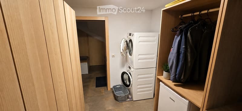 A small, modern laundry room with a stacked washing machine and dryer, a built-in wardrobe with hanging clothes, a small refrigerator, and a laundry basket on the floor.