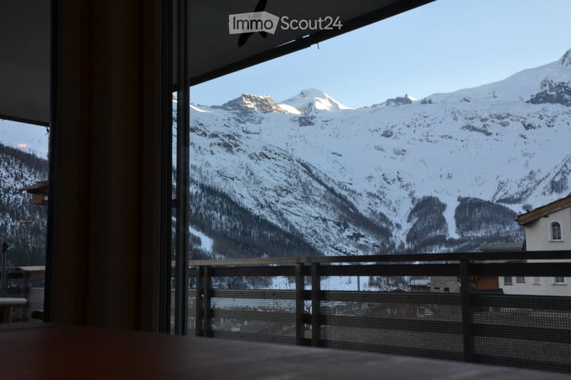 Balcony with a wooden railing overlooking a snow-covered mountain range and a small village below.