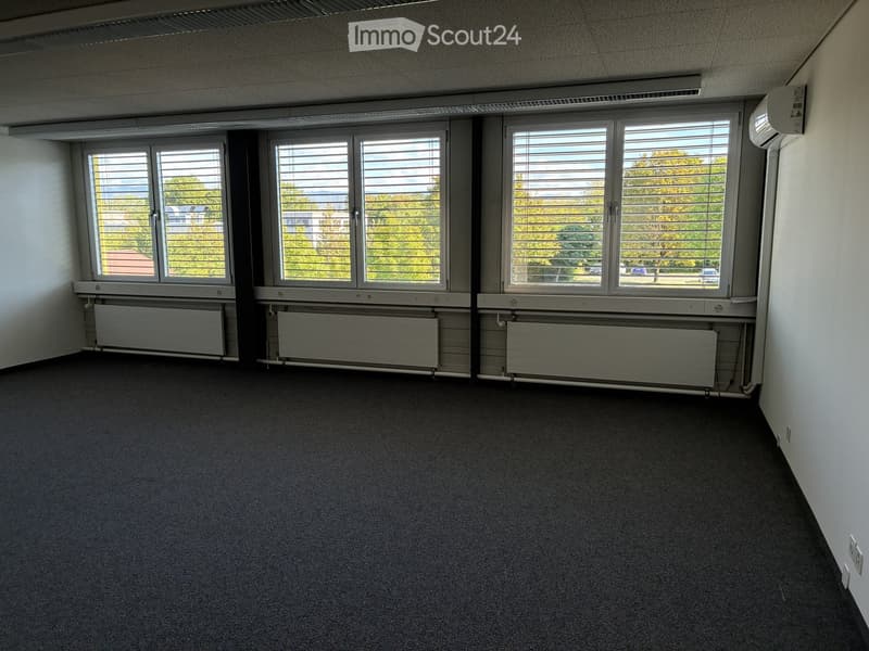 Empty room with white walls, carpeted floor, large windows with blinds, radiators, and an air conditioning unit on the wall.