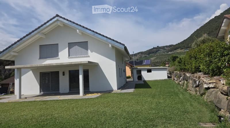 Modern two-story house with white exterior, garage, and a well-maintained lawn in front. The house features large windows and a covered entrance area. The surrounding area includes a stone wall and a scenic hillside in the background.
