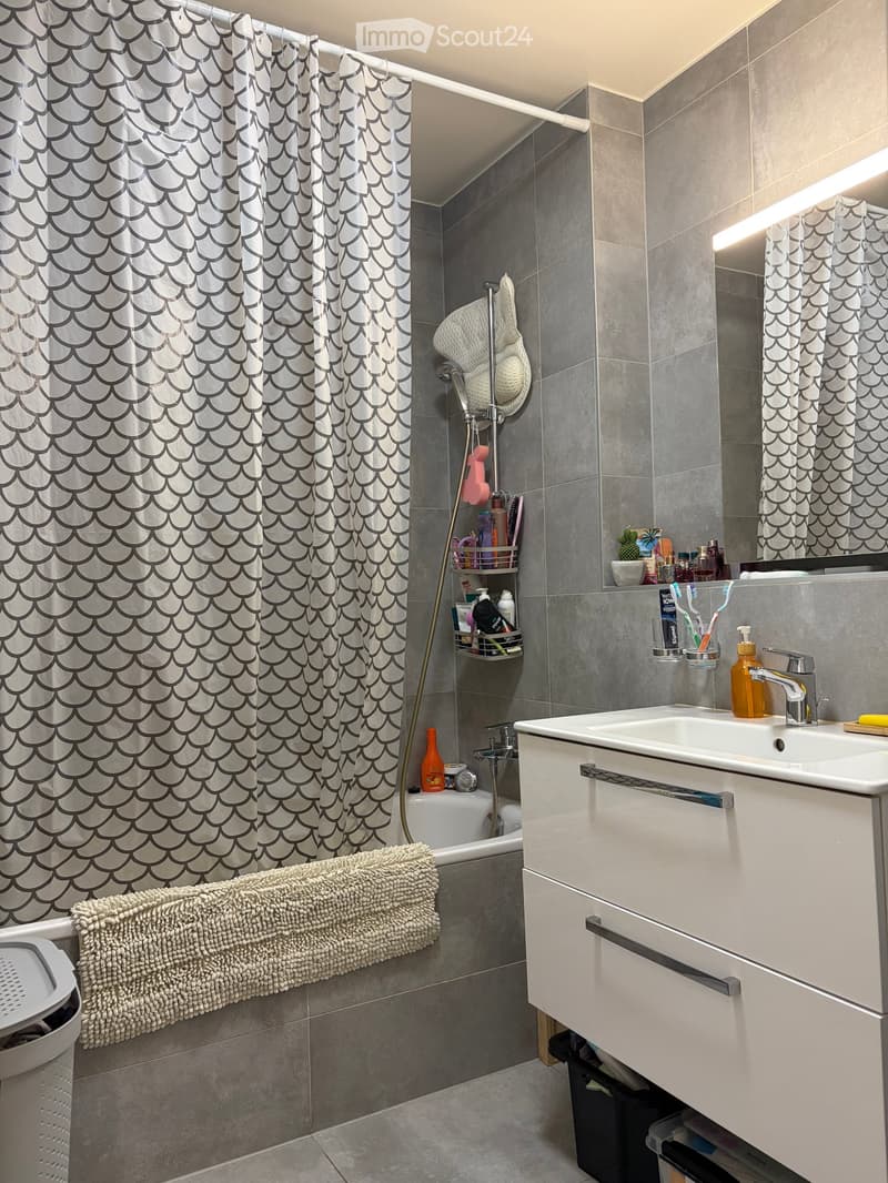 Modern bathroom with a white sink, mirror, bathtub, and shower area. Features a decorative towel, shower curtain, and various toiletries on the sink and shower rack.
