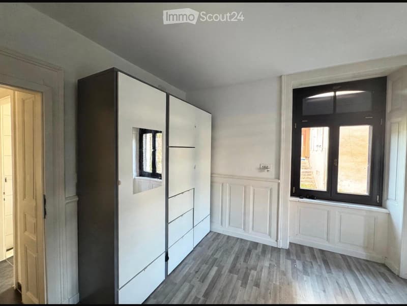 A modern, unfurnished room with a large white wardrobe, wooden flooring, and large windows with black frames. The walls are painted white with decorative paneling near the floor.