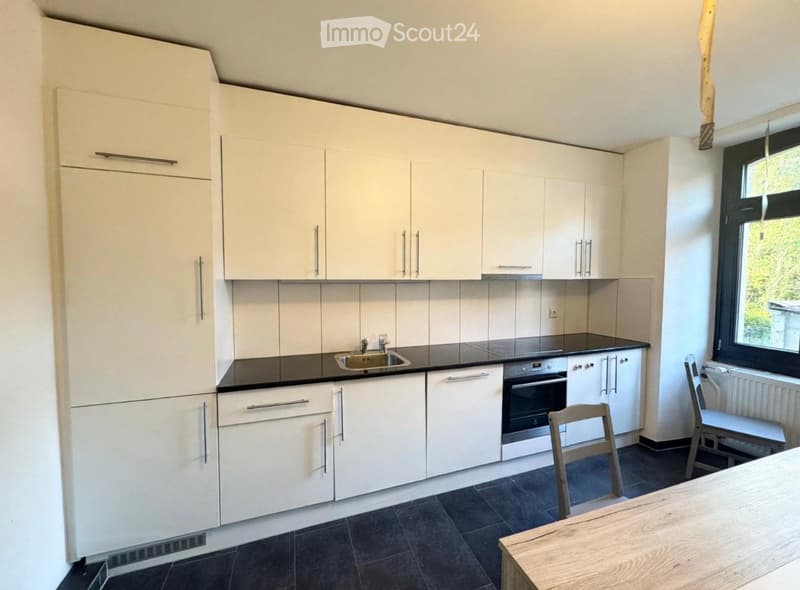 Modern kitchen with white cabinets, black countertops, stainless steel sink, and built-in oven. Includes a wooden dining table and chairs near a large window.