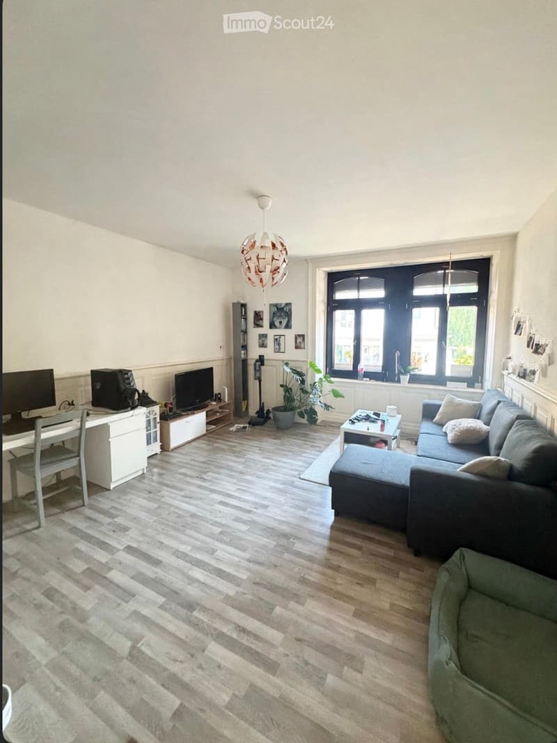 Living room with a grey sectional sofa, coffee table, desk with chair, TV, and a hanging lamp. Wooden flooring and large windows.