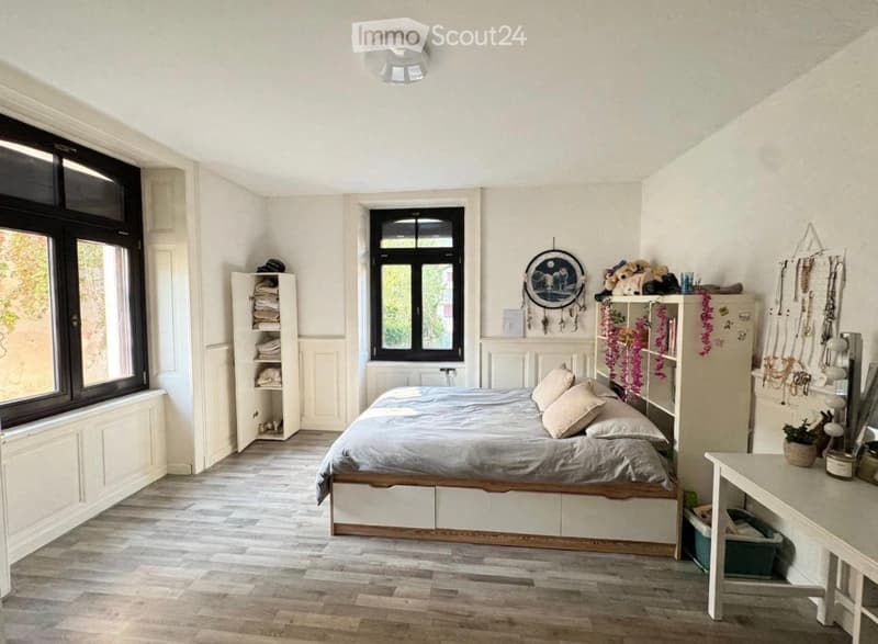 Cozy bedroom with wooden flooring, large windows, a bed with pillows and blankets, a nightstand, a mirror, a shelf, and a small table with a plant.