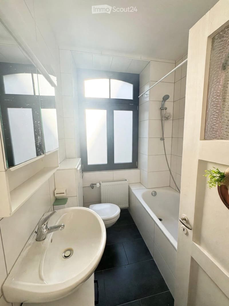 Modern bathroom with a sink, toilet, bathtub, and a window with frosted glass panes. The room features white tiles, a radiator, and a mirror above the sink.