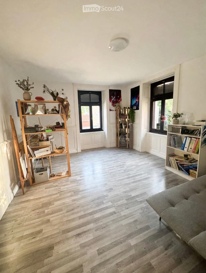 Bright living room with wooden flooring, large windows, and modern decor including a sofa, shelves with books and decorative items, and artwork on the walls.