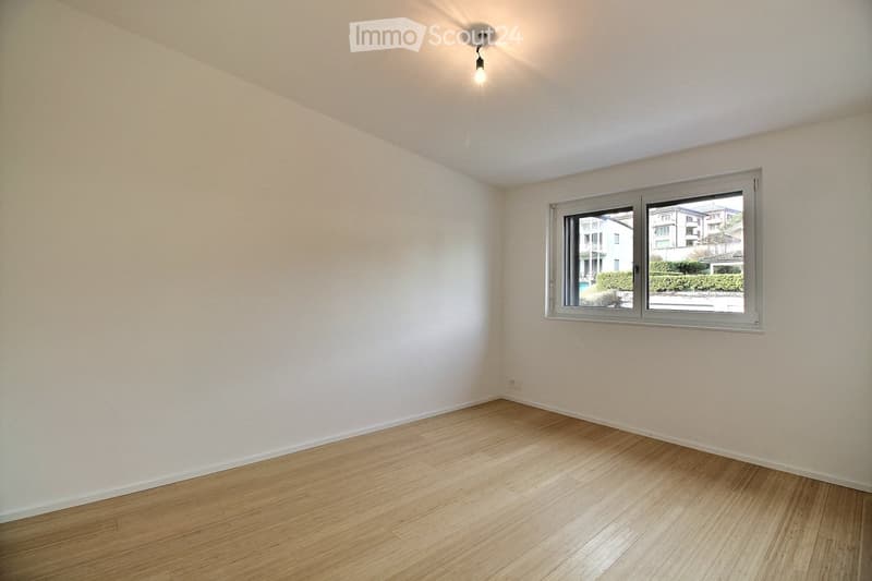 Empty room with light wooden flooring and white walls, featuring a single window with a view of neighboring buildings and greenery.