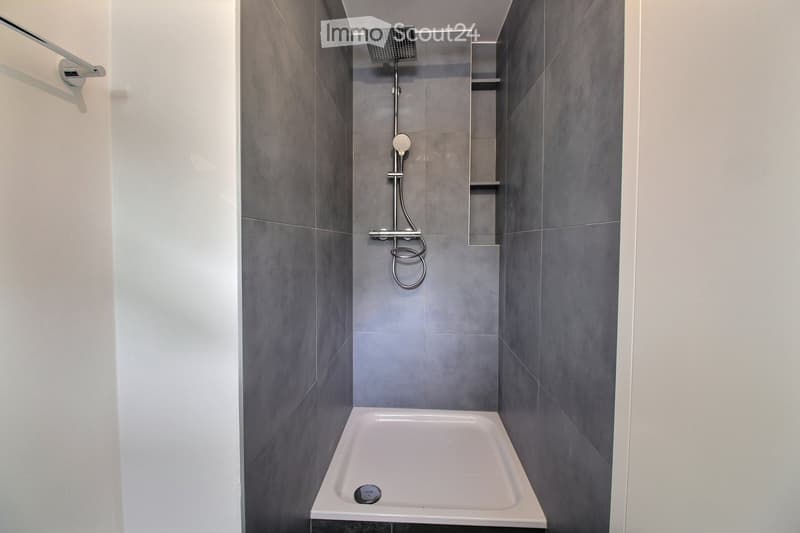 Modern bathroom with a white bathtub, grey tiled walls, and a built-in shelf. Features a showerhead and hose mounted on the wall.