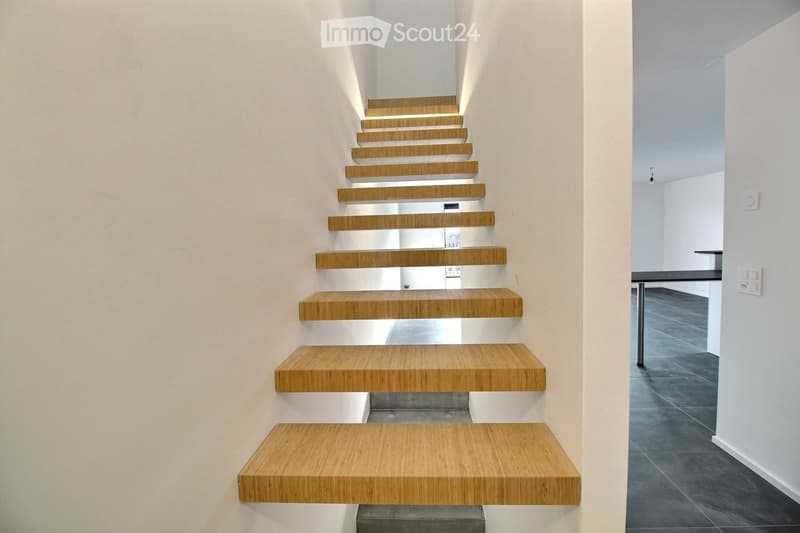 Modern hallway with a wooden staircase featuring minimalist design and sleek steps, leading to an open area with dark tiled flooring and white walls.
