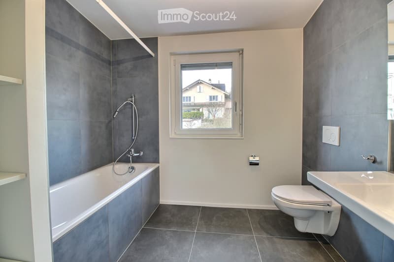 Modern bathroom with grey tiled walls, a bathtub, toilet, sink, and a window offering a view of the exterior.