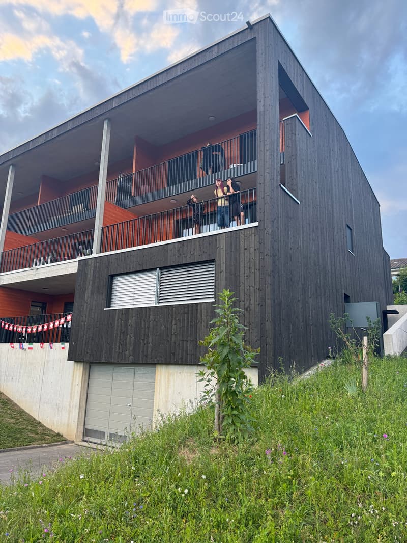 Modern three-story building with dark wooden facade, balconies, and a raised ground floor. Features include a garage entrance and decorative flags along the lower level.