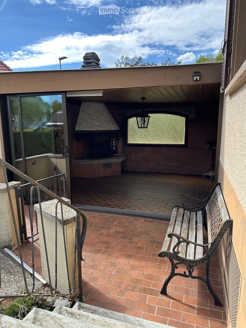 Terrace with brick tiles, a bench, and a view of a covered outdoor area with a fireplace and glass walls.