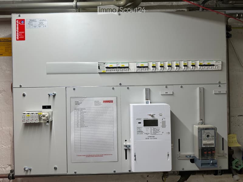 Electrical panel with multiple switches, meters, and a list of circuit breakers, mounted on a wall with visible wiring and pipes.