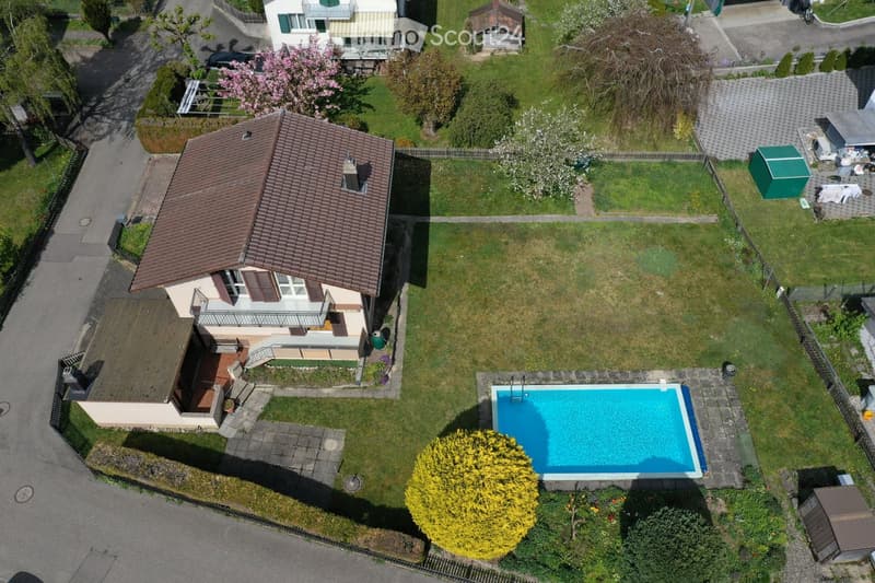 Aerial view of a two-story house with a brown tiled roof, surrounded by a well-maintained garden, featuring a swimming pool, trees, and a small shed.