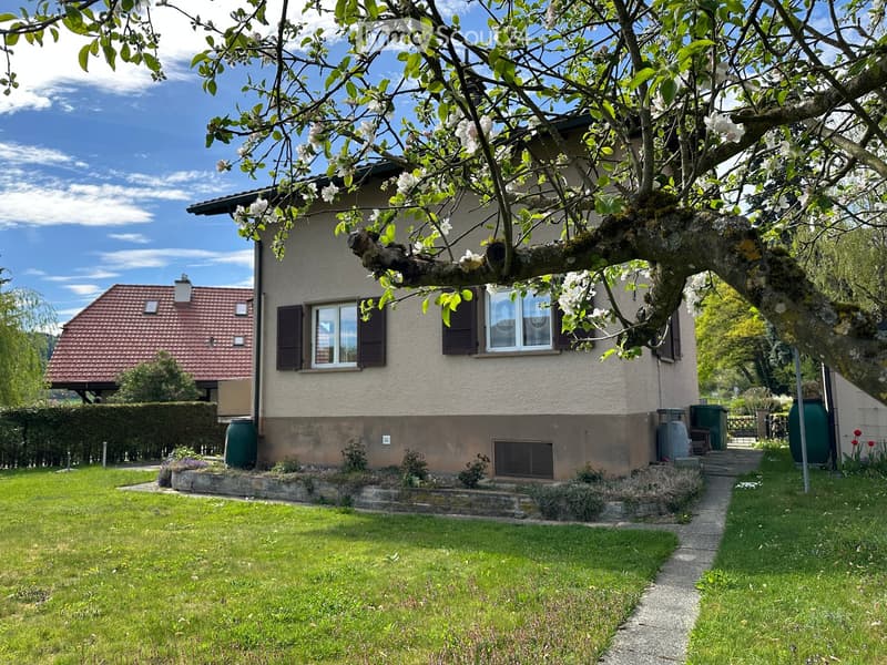 Two-story house with a red-tiled roof, beige siding, and multiple windows, surrounded by a well-maintained lawn and garden with a blooming tree in the foreground.
