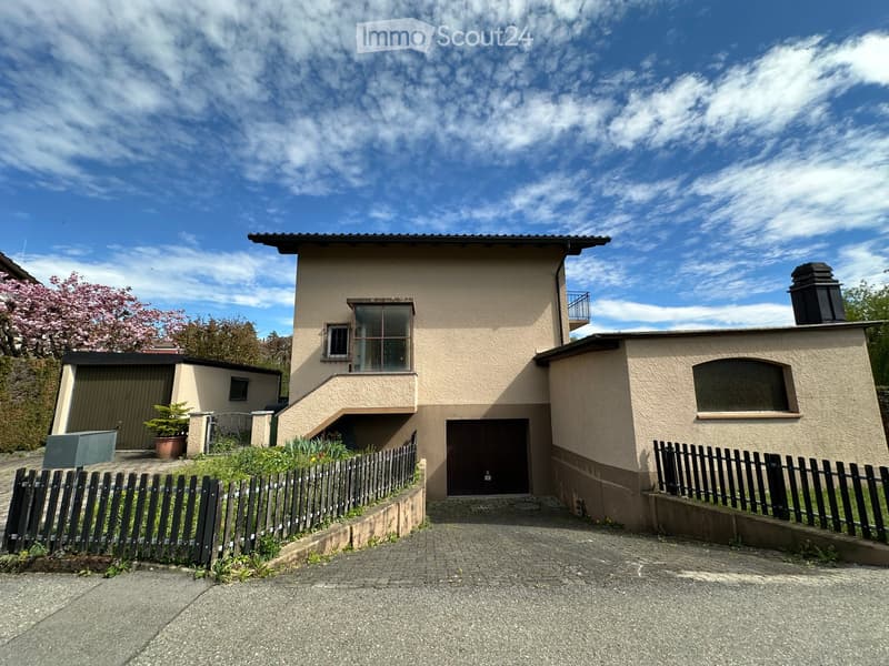Two-story house with beige facade, large windows, garage, and small garden area with a potted plant and fence.