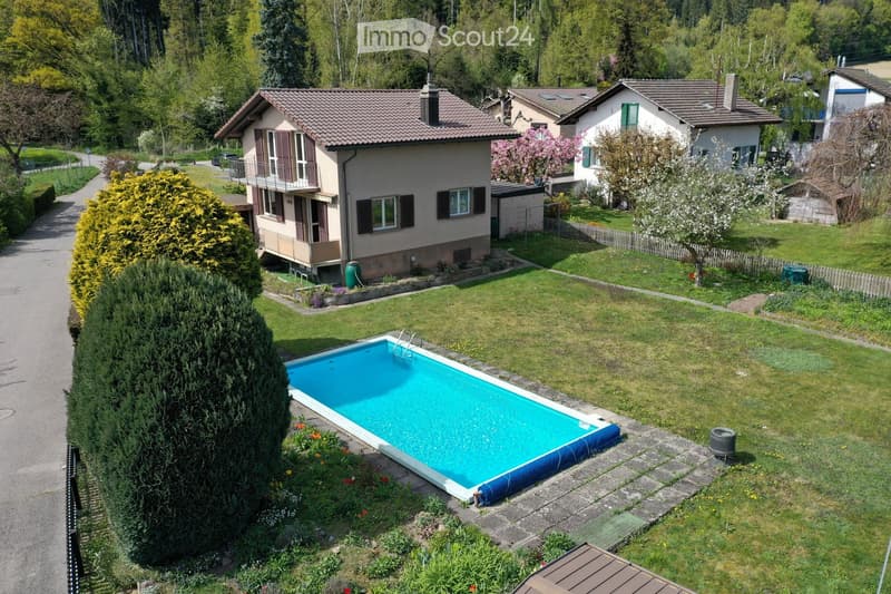 Aerial view of a residential area with a swimming pool in the foreground, surrounded by a well-maintained lawn and garden. The pool is rectangular with a blue interior and a white border. Nearby, there is a tall, conical evergreen tree and a variety of fl