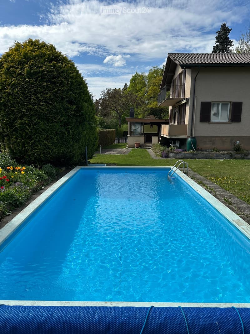 A rectangular swimming pool with clear blue water, surrounded by a well-maintained garden with flowers, shrubs, and a house with a balcony in the background.