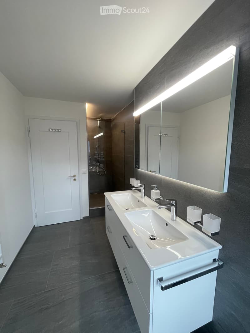 modern bathroom with grey tiles, large illuminated mirror, two sinks, and shower behind frosted glass door