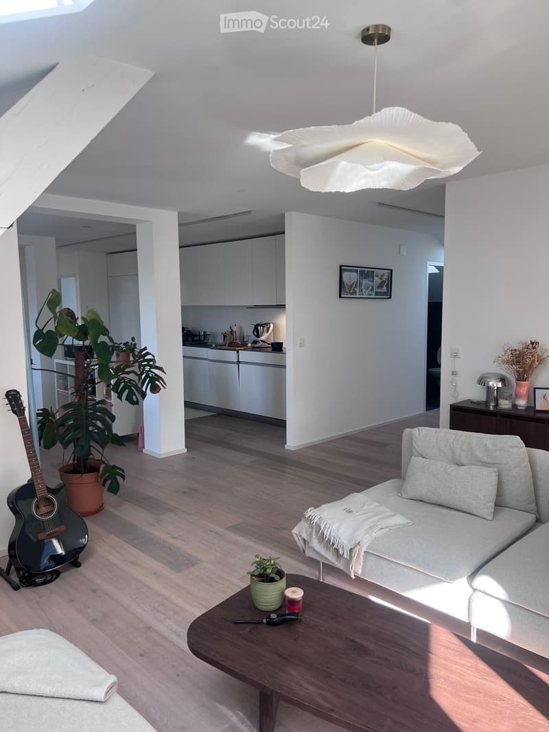 Bright living room with wooden flooring, white walls, and a skylight. Features a grey sofa with a throw blanket, a wooden coffee table with a small plant and candle, a guitar leaning against the wall, and a kitchen area with white cabinets and appliances.