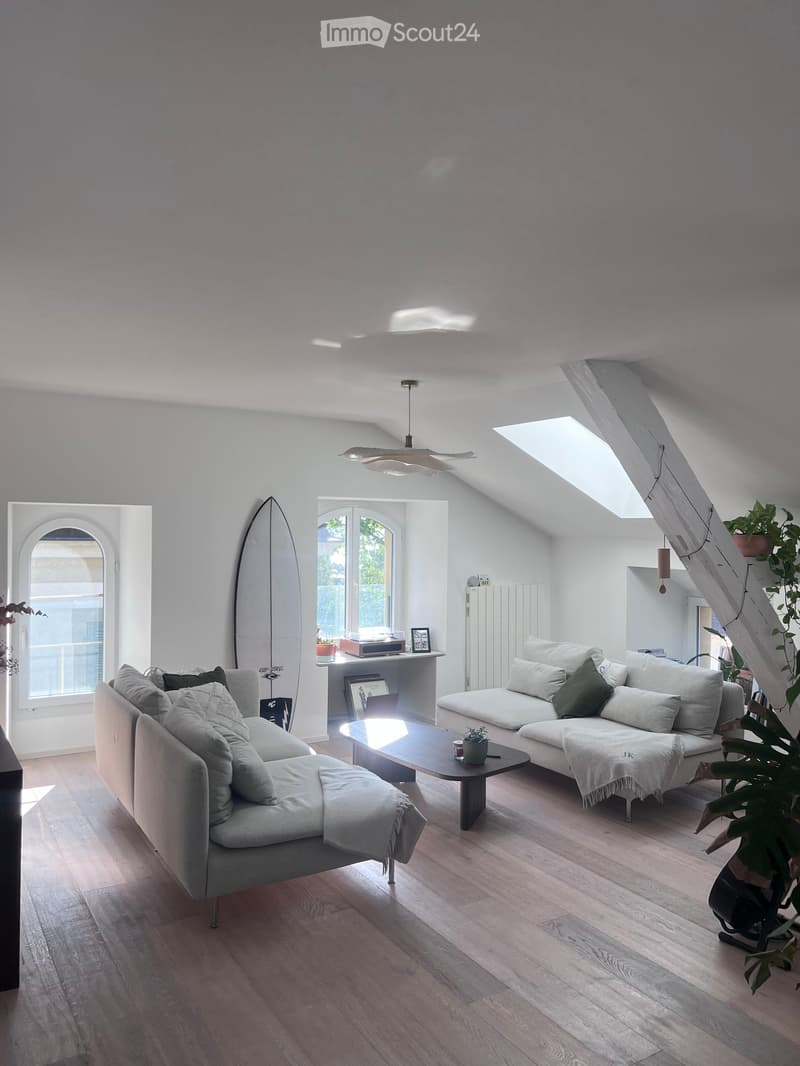 Bright living room with wooden flooring, white walls, and large windows. Features a grey sectional sofa, coffee table, laptop on a shelf, and a surfboard leaning against the wall.