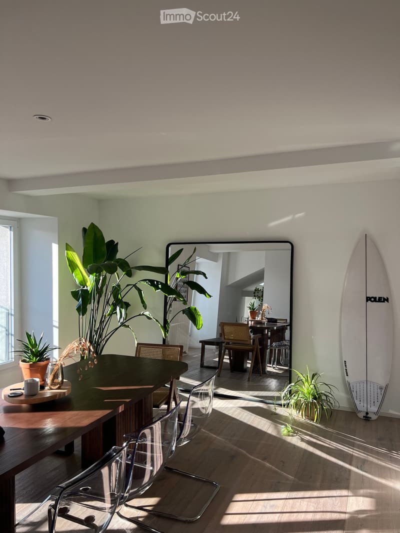 Modern living space with wooden flooring, large mirror, surfboard leaning against wall, and several potted plants. A small round table with a cup and vase is visible, along with clear plastic chairs.