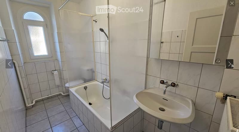 Bathroom with a bathtub, sink, mirror, and shower area. The room features tiled walls and floor, with a window providing natural light.