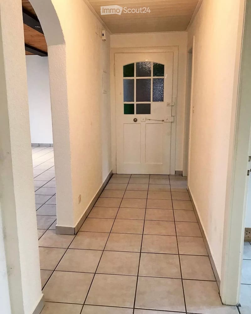 A clean, tiled hallway with white walls and a central white door featuring a glass panel. The hallway has a simple, modern design with a visible column on the left side.